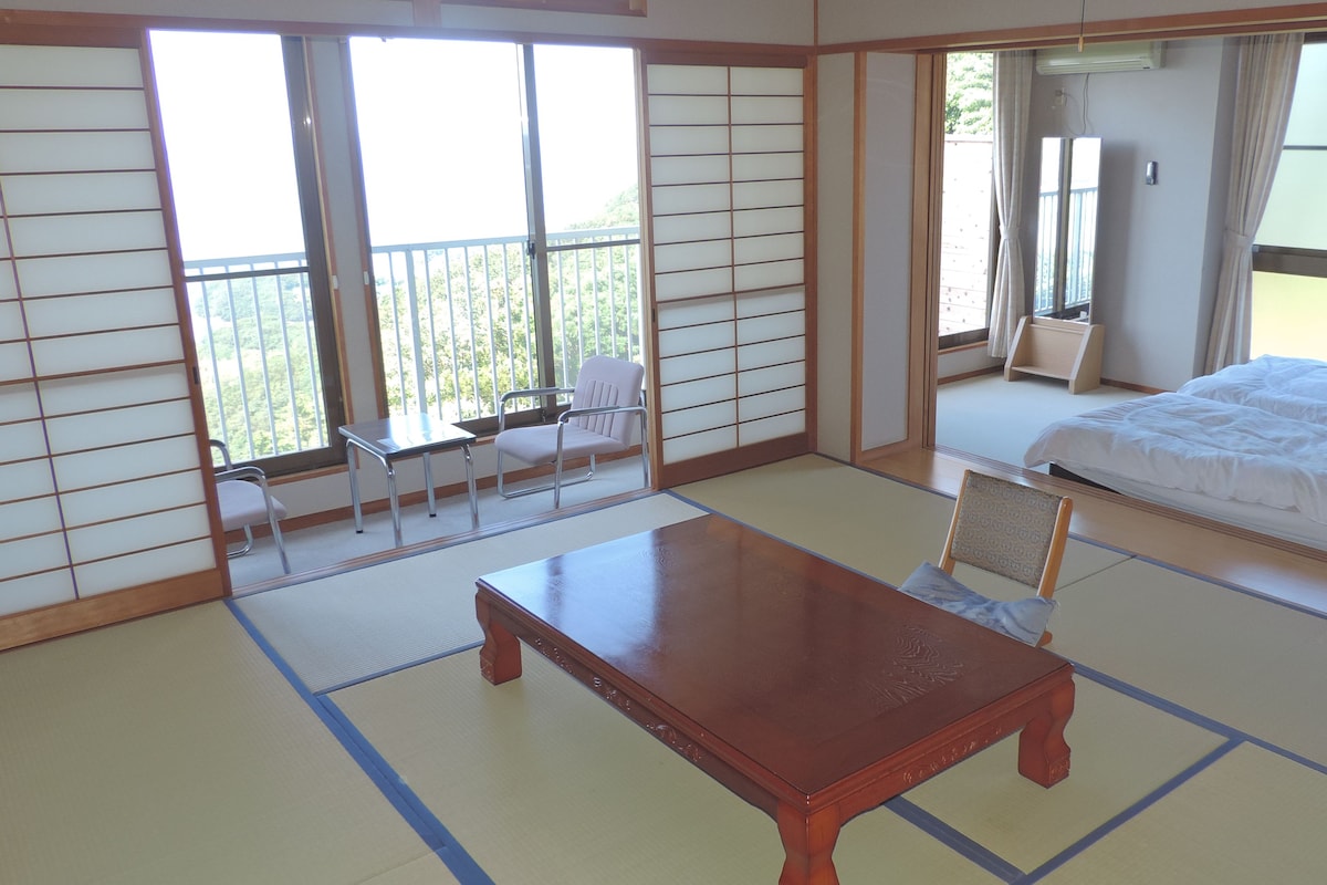 A traditional Japanese room with tatami flooring, a wooden table, a bed with white bedding, and a sliding glass door leading to a balcony with a view of greenery.