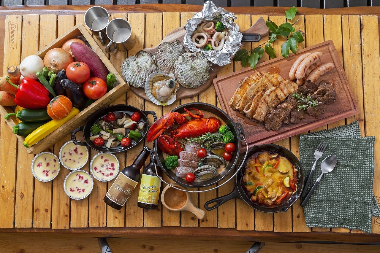 Una mesa de madera con diversos alimentos, como mariscos, verduras y carnes, además de bebidas y una sartén.