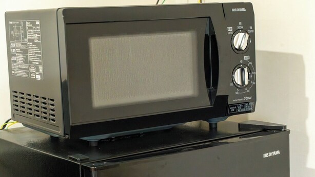 A black microwave oven with a digital display and control knobs on top of a black refrigerator. - Shimairo Nagate (Goto)