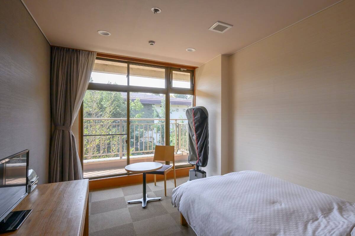 A hotel room with a bed, a desk with a computer, a chair, and a window with a view of a balcony.