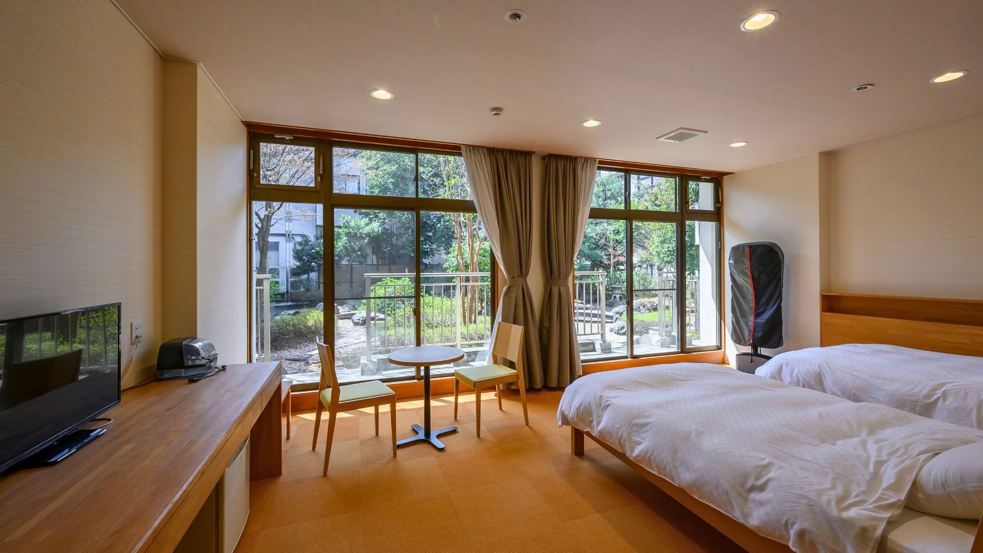 Une chambre d’hôtel avec deux lits, une télévision, un bureau, une chaise et une vue sur un jardin grâce à de grandes fenêtres.