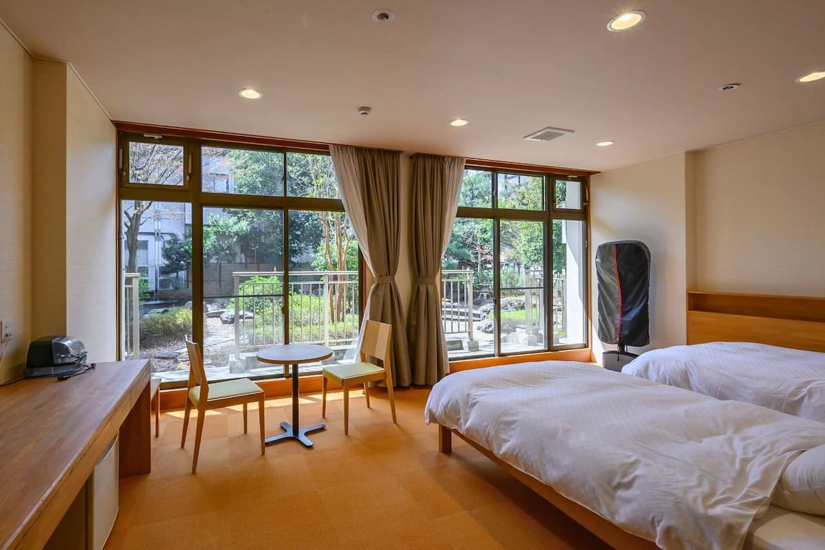 A hotel room with two beds, a TV, a desk, a chair, and a view of a garden through large windows.