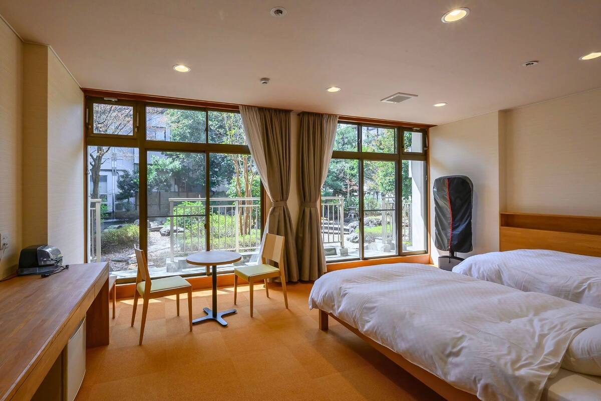 A hotel room with a bed, a TV, a desk, a chair, and a view of a garden through large windows.