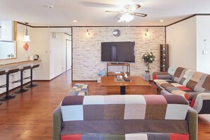 Una sala de estar moderna con una mesa de centro de madera, un televisor de pantalla plana grande montado en una pared de piedra, un sofá y un comedor con una mesa de madera y taburetes de bar.