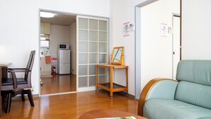 Een woonkamer met houten vloer, een groene bank, een houten stoel, een kleine tafel, en een schuifdeur naar een keuken.
