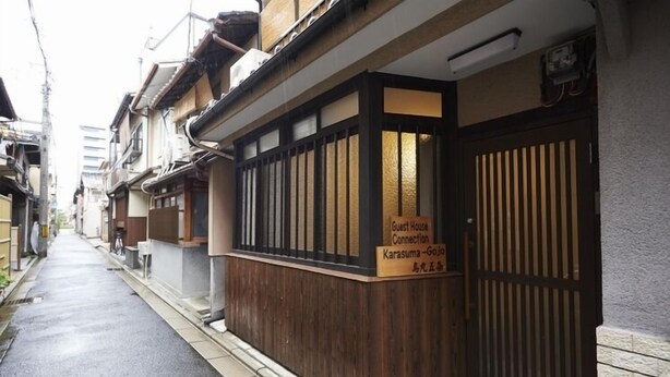 A narrow street with traditional Japanese buildings, a wooden door with a sign indicating a guest house, and a small wooden structure on the sidewalk. - Guesthouse Connections Karasuma Gojo (Kyoto)