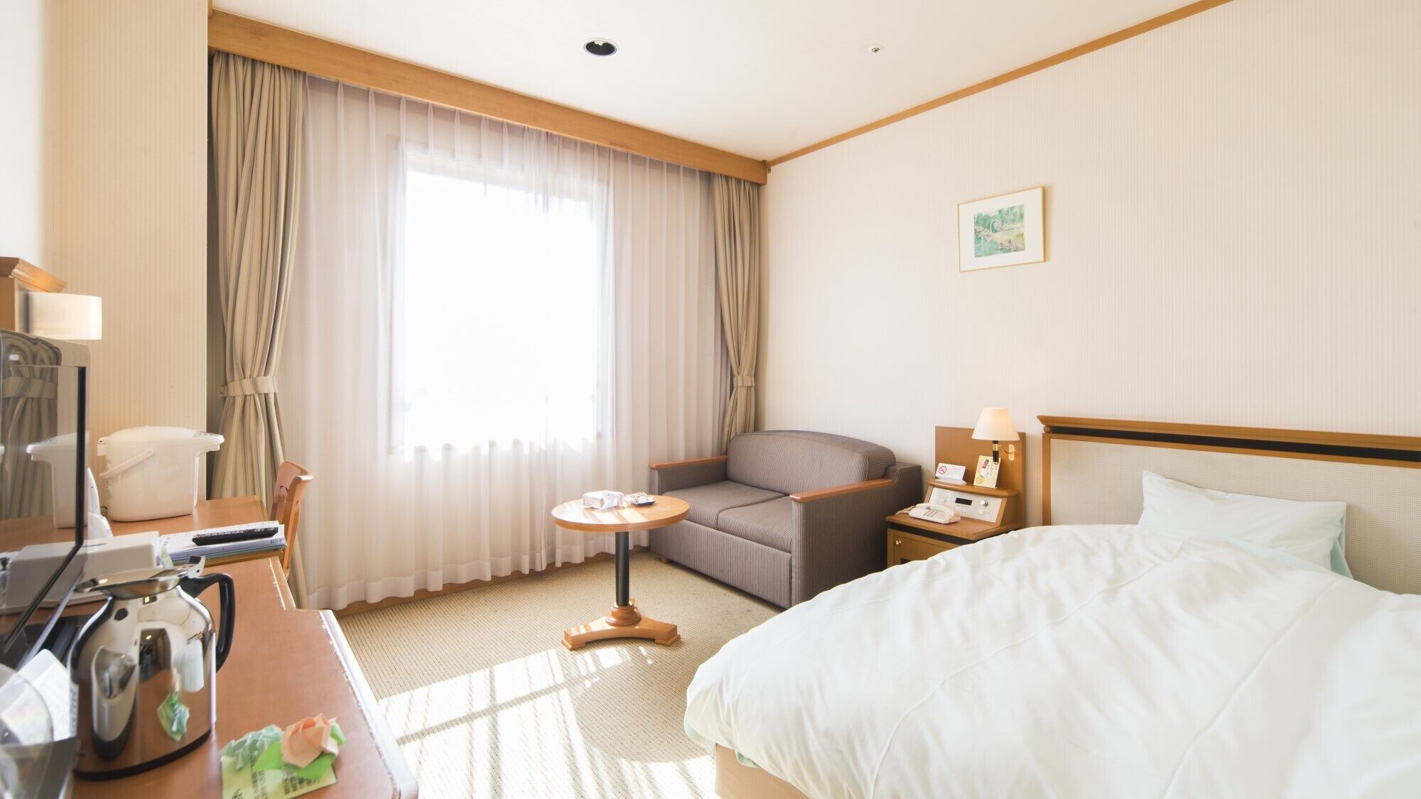 Ein Hotelzimmer mit einem Bett, einer Schlafcouch, einem kleinen Tisch, einem Schreibtisch mit Wasserkocher und Vorhängen.