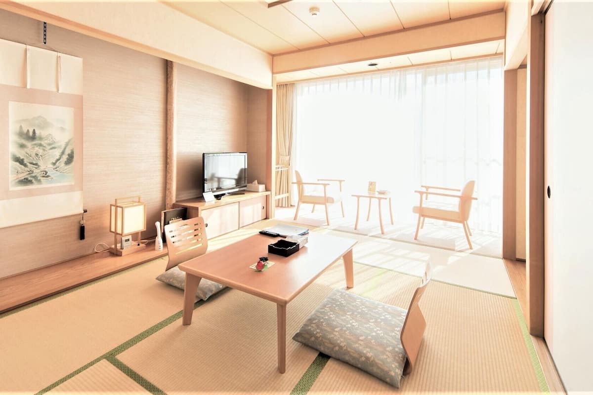 A modern hotel room with a wooden floor, a dining table, a sofa, a TV, and a large window.