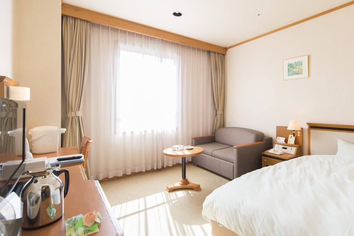 A hotel room with a bed, a sofa, a small table, a desk with a kettle, a telephone, and curtains.