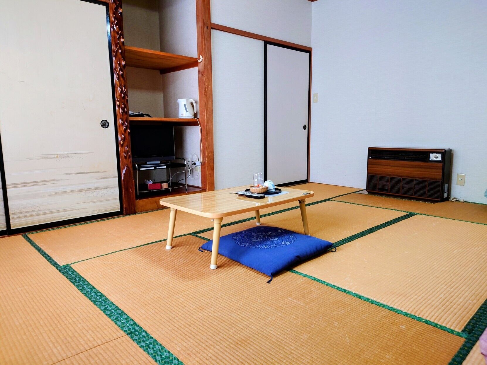 A traditional Japanese room with tatami flooring, a low wooden table, a blue cushion, a television, and a heater.