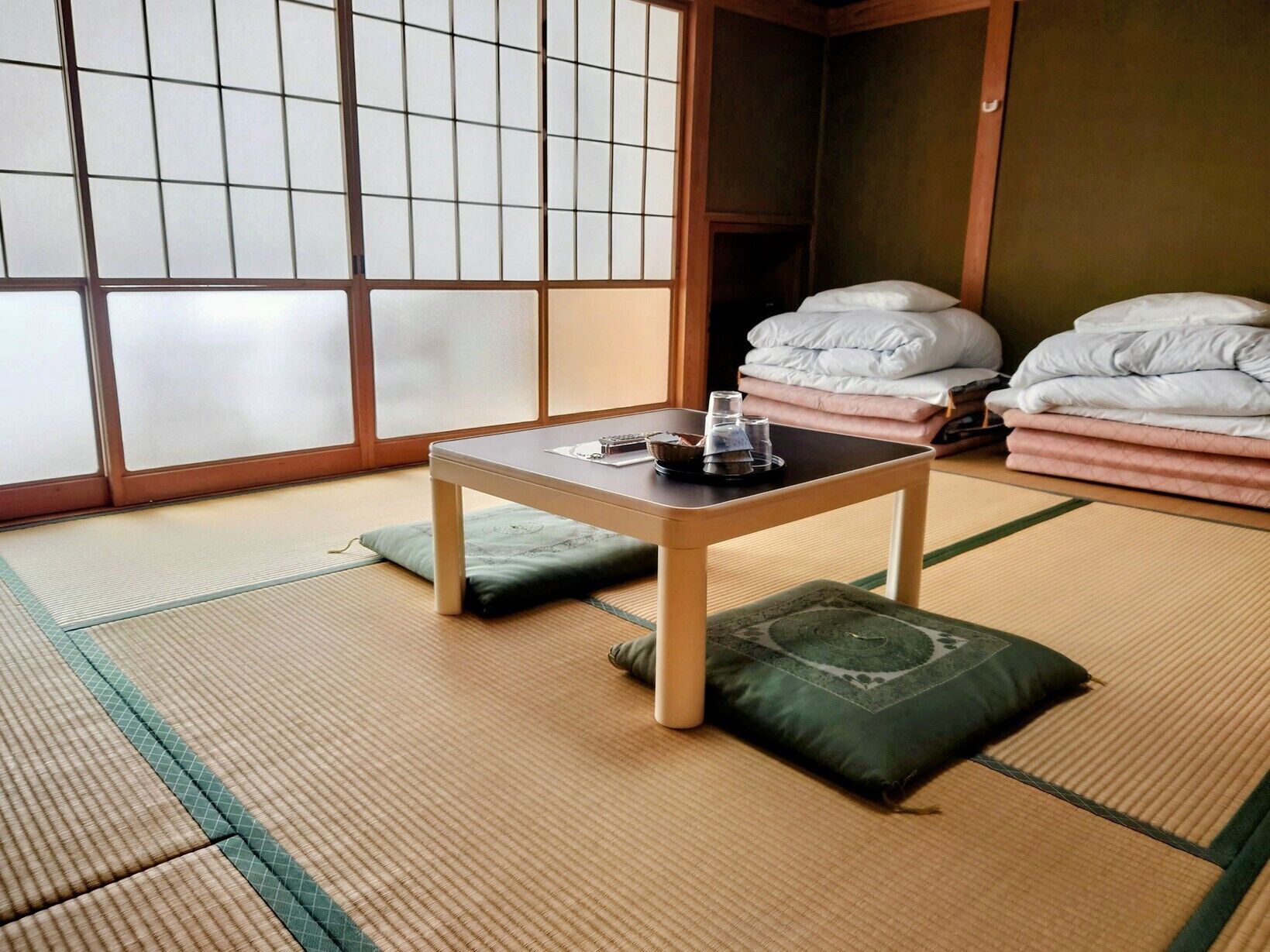 Une pièce avec un sol recouvert de tatamis, une petite table basse, des coussins et une pile de couvertures.