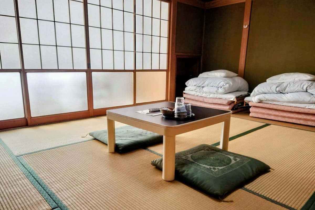 Une pièce avec un sol recouvert de tatamis, une petite table basse, des coussins et une pile de couvertures.