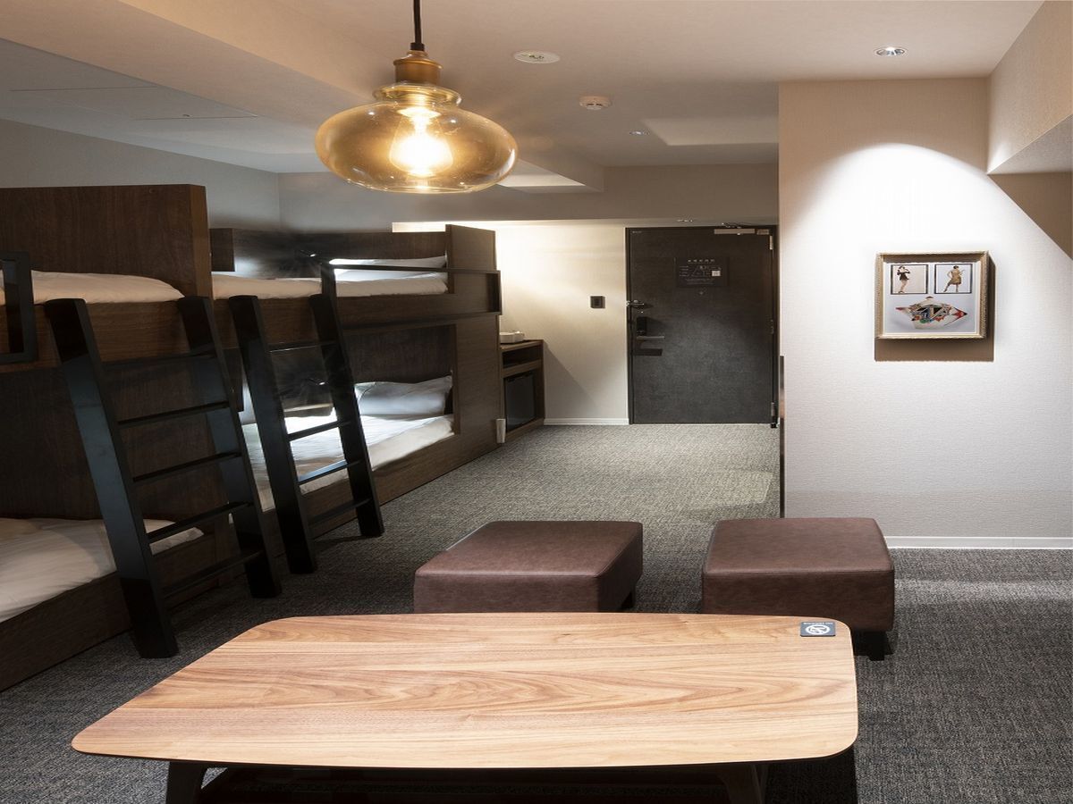 A bunk bed room with a table and ottomans, a hanging light fixture, and a door with a control panel.