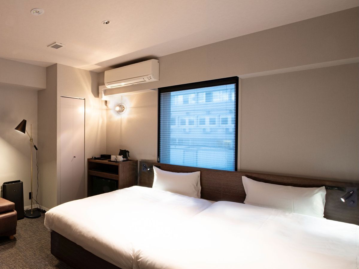 A hotel room with a large bed, a desk, a lamp, an air conditioner, and a window with a city view.