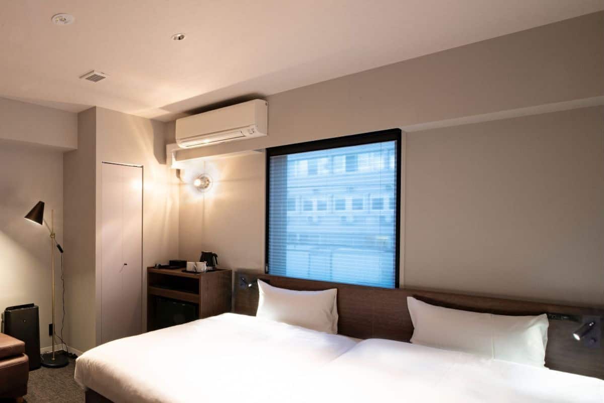 A hotel room with a large bed, a desk, a lamp, an air conditioner, and a window with a city view.