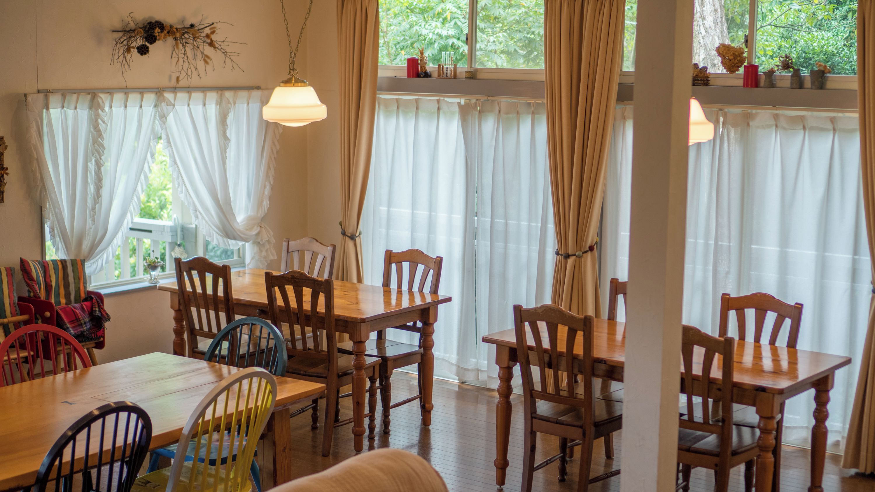 A dining area with a wooden table and chairs, white curtains, and a view of greenery outside.