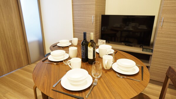A dining table set with white plates, bowls, and cutlery, accompanied by wine bottles and glasses, in a room with a TV and wooden paneling. - Prime Suites Tokyo (Tokyo)