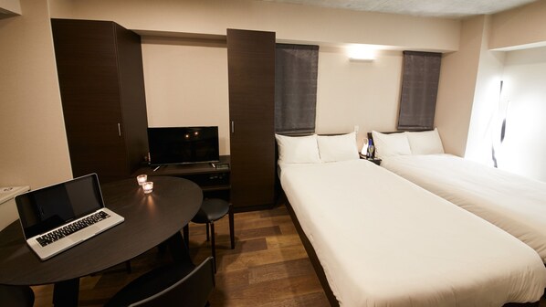 A hotel room with a bed, a desk with a laptop, a TV, and a bedside table with a bottle and glasses.