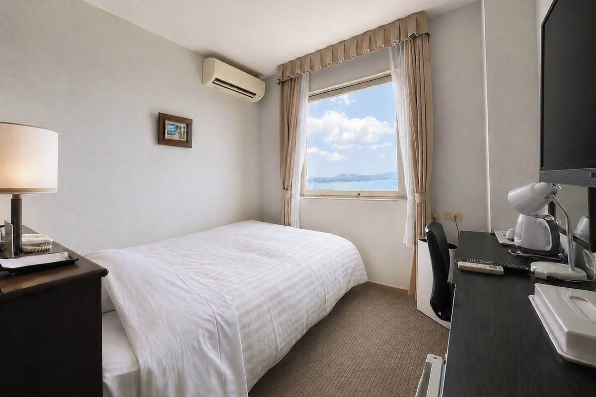 A hotel room with a bed, a desk with a lamp, a TV, and a window with curtains.