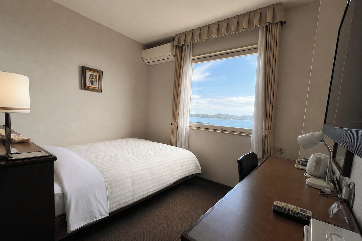 Ein Hotelzimmer mit Bett, Schreibtisch, Stuhl, Lampe, Fernbedienung und einem Fenster mit Vorhängen.