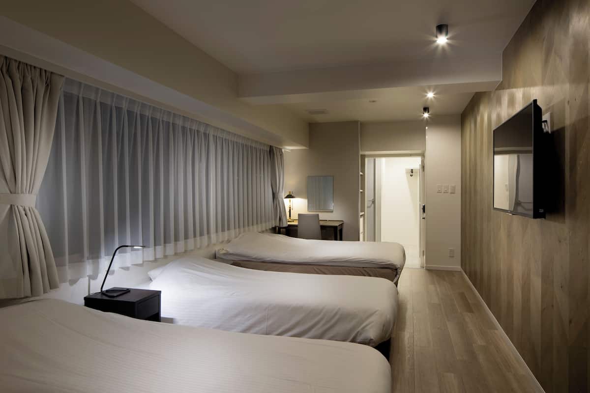 A hotel room with two beds, a desk, a chair, a TV, and a large window with curtains.