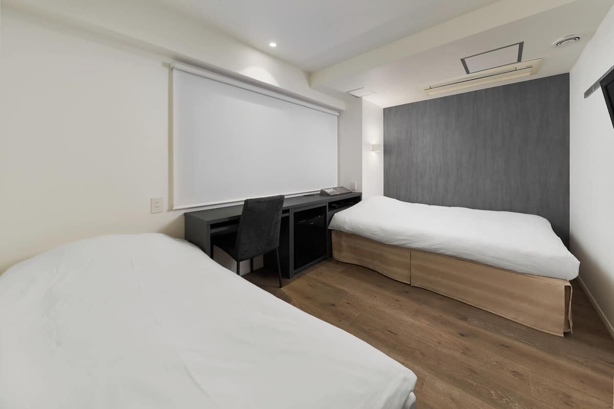 A modern hotel room with two single beds, a desk, a chair, and a large window.