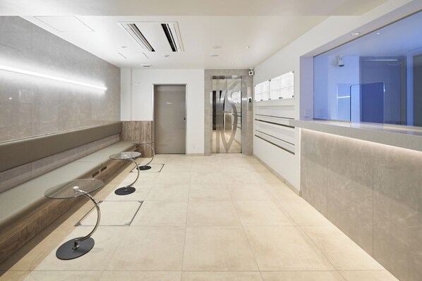 A modern, well-lit hallway with a reception desk, a glass elevator, and a bench with a glass table. - Shinjuku City Hotel N. U. T. S Tokyo (Tokyo)