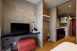 Ein kleines Hotelzimmer mit Flachbildfernseher, einem kleinen Schreibtisch mit Stuhl, einer roten Bank, einer Waschmaschine und einer roten Tür.