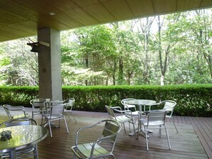 An outdoor patio area with metal tables and chairs, surrounded by a wooden deck and a hedge. - I&I Land (Shijonawate)