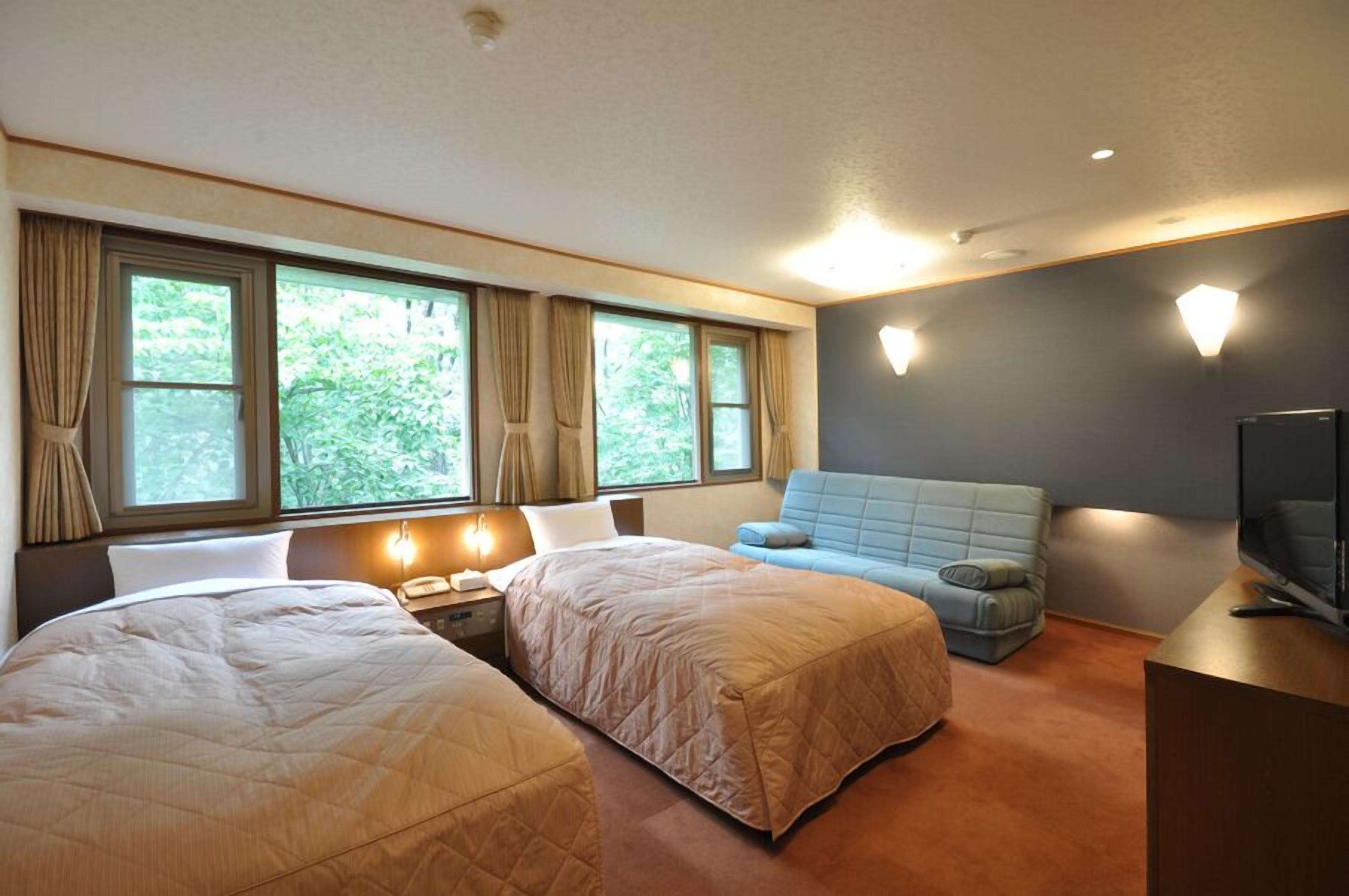 A hotel room with two beds, a sofa, a TV, and large windows with curtains.
