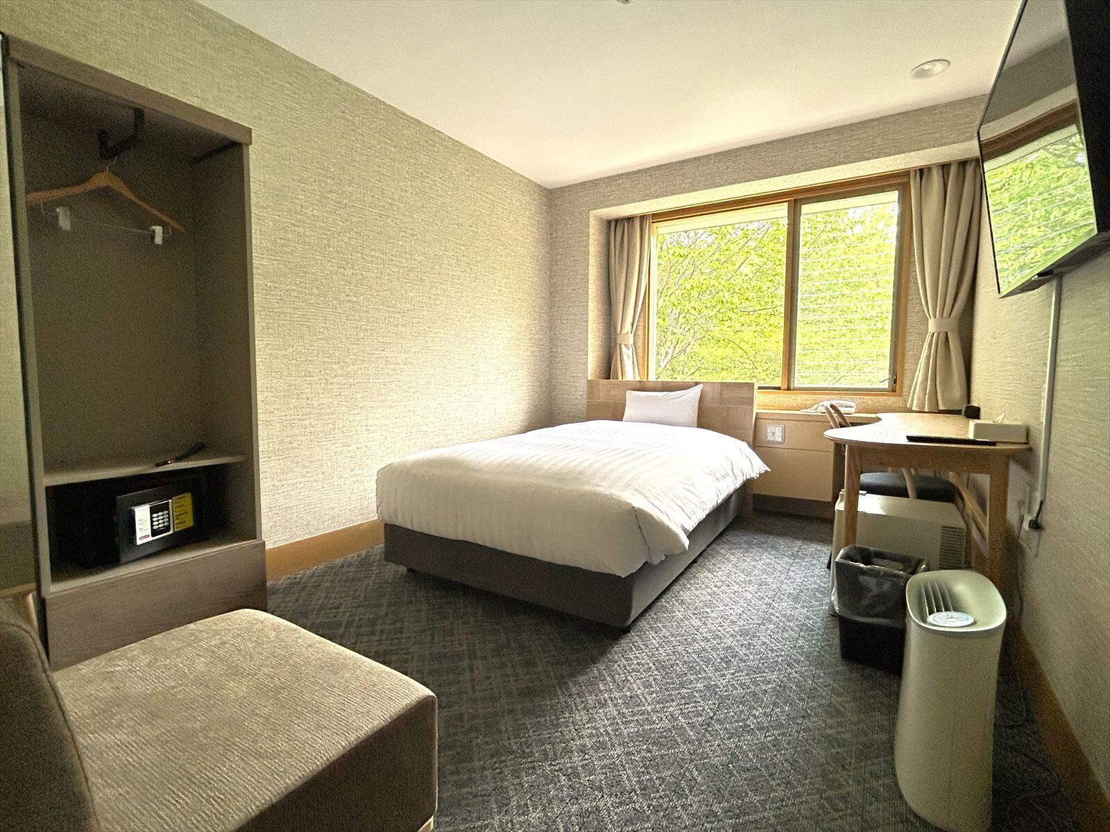 A hotel room with a bed, a desk, a chair, a TV, a window with curtains, and a coat rack.