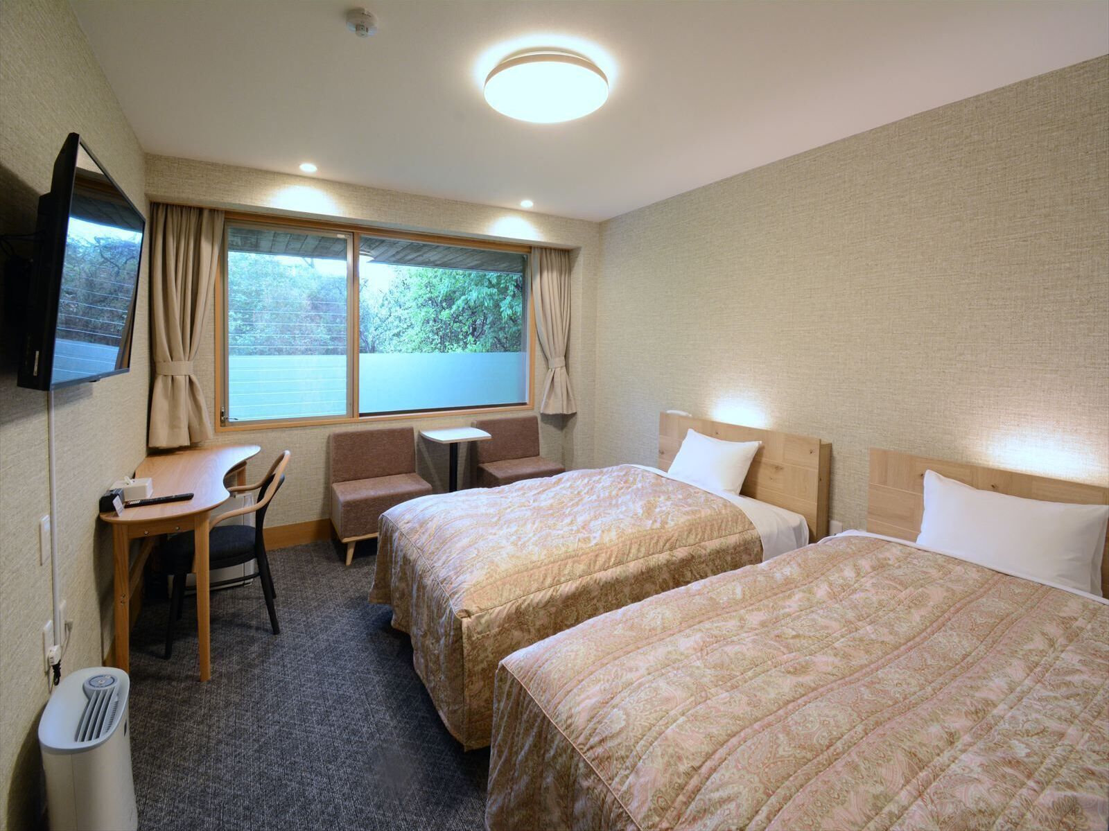 A hotel room with two beds, a desk, a chair, a small table, a TV, and a window with curtains.