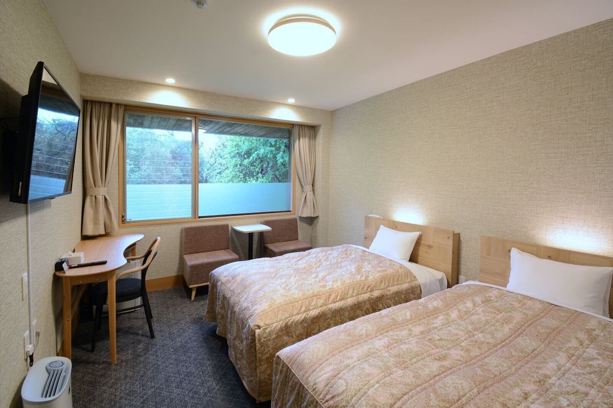 A hotel room with two beds, a desk, a chair, a small table, a TV, and a window with curtains.