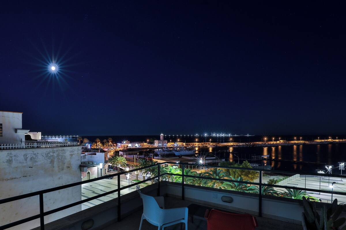 A balcony with a view of a marina at night, featuring a calm sea, boats, and a cityscape with illuminated buildings.