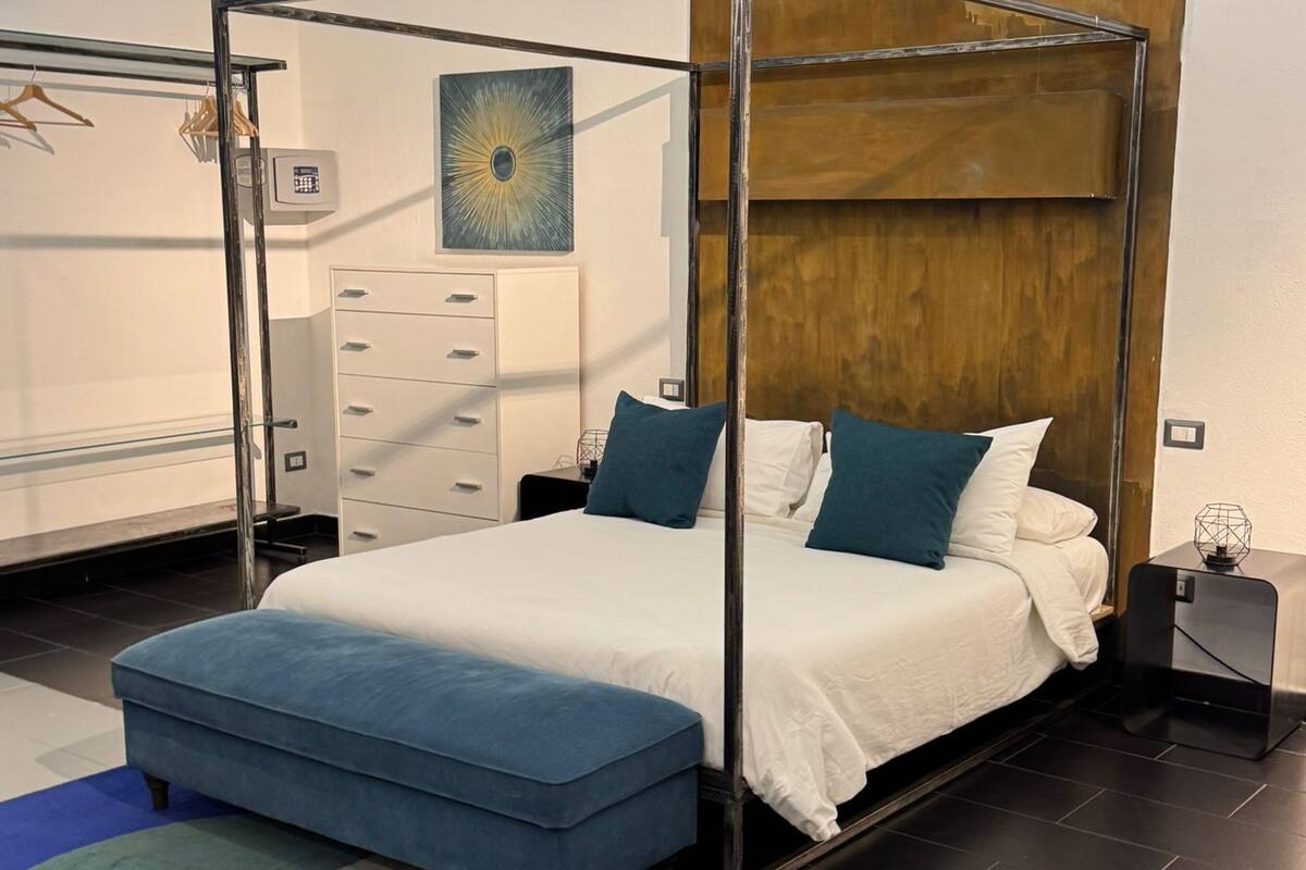 A modern bedroom with a four-poster bed, a chest of drawers, a wardrobe, and a large abstract painting on the wall.