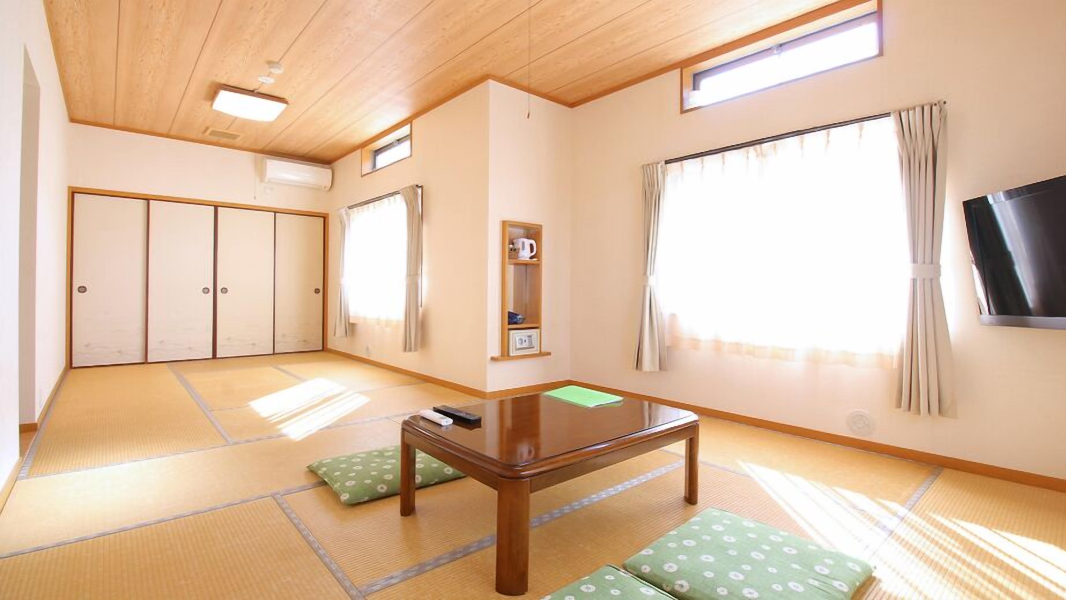 A room with tatami flooring, a low wooden table, green cushions, a TV, sliding doors, and a shelf with a kettle.