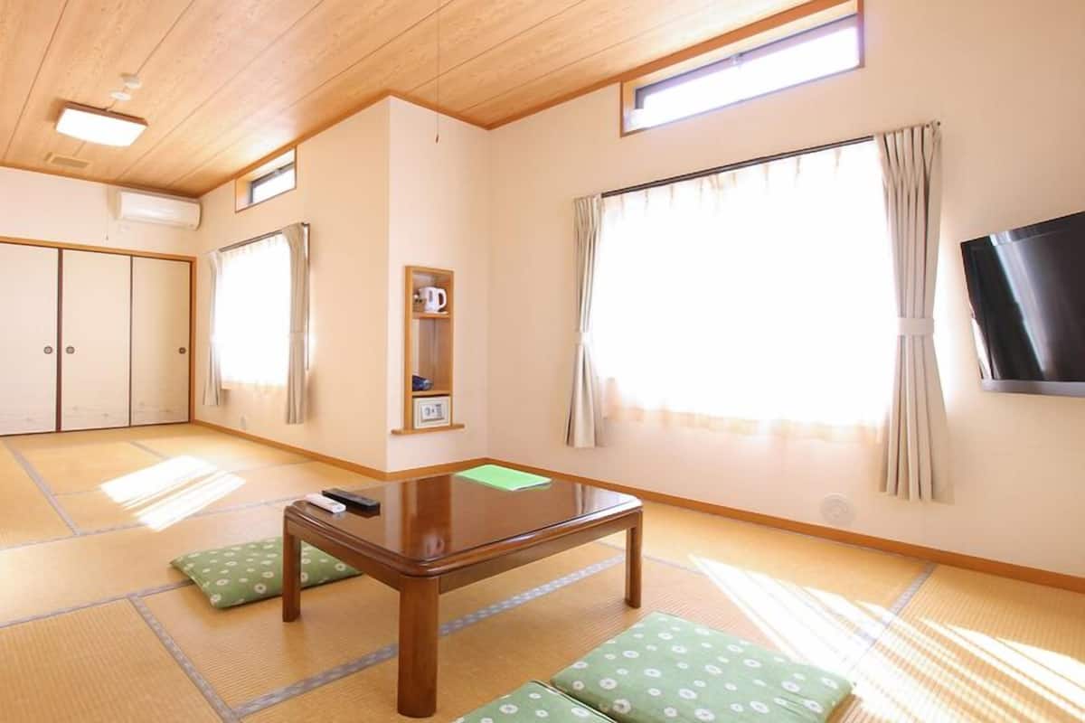 A room with tatami flooring, a low wooden table, green cushions, a TV, sliding doors, and a shelf with a kettle.
