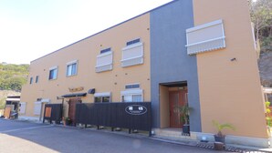 Un edificio de dos pisos con fachada color beige y gris, múltiples ventanas y una cerca negra con un letrero.