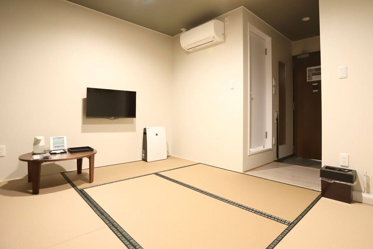 A room with tatami flooring, a flat-screen TV mounted on the wall, a small table with a remote control, and a heater.