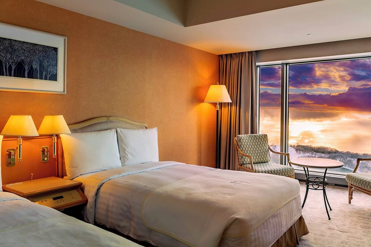 Une chambre d’hôtel avec un lit, des lampes de chevet, une chaise, une petite table et une grande fenêtre donnant sur un paysage au coucher du soleil.