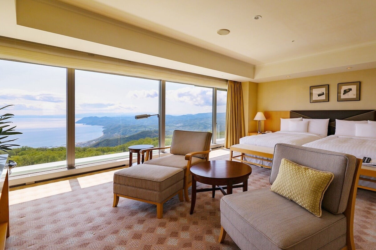 Une chambre d’hôtel avec une grande fenêtre, un lit, une chaise, une petite table et une table de chevet.