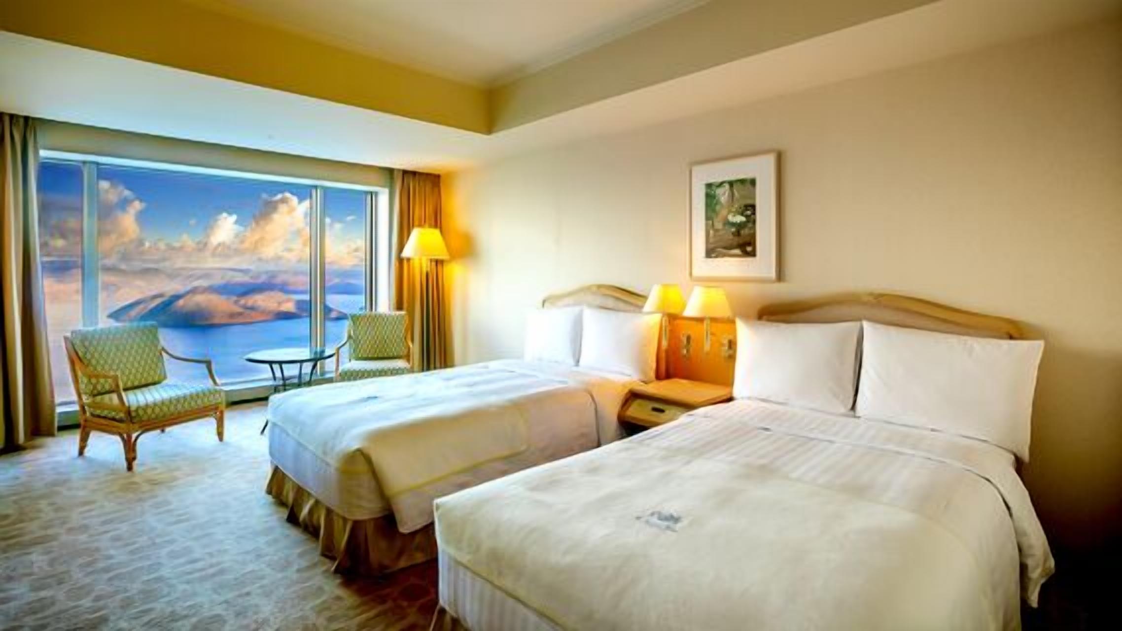 Ein Hotelzimmer mit zwei Betten, einem Sessel, einem Tisch und einem großen Fenster mit Blick.