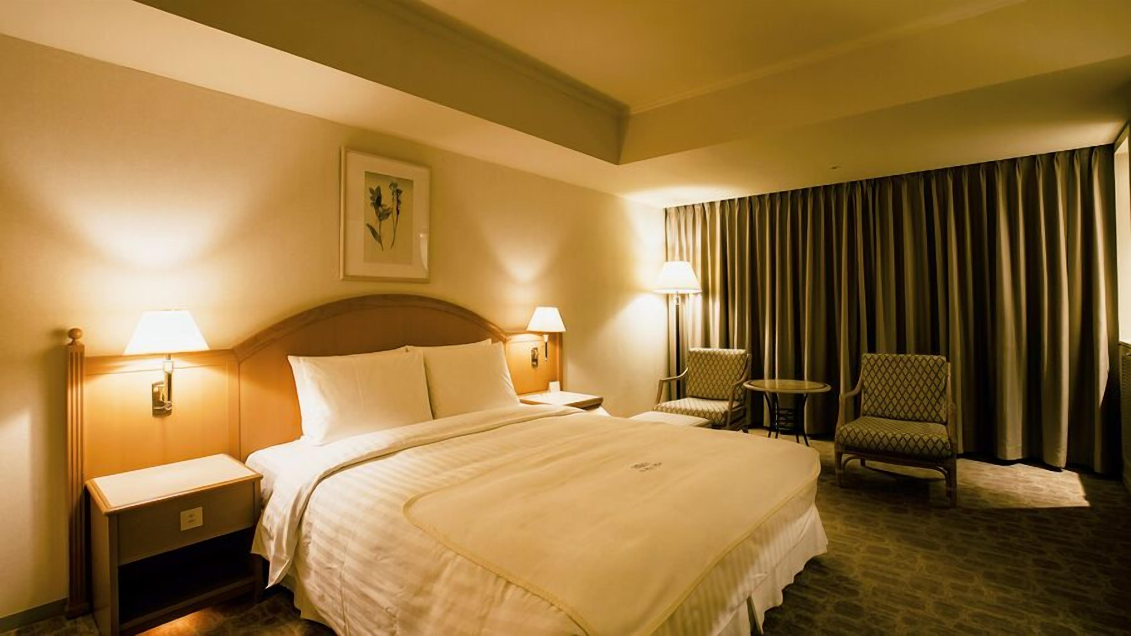 Ein Hotelzimmer mit einem großen Bett, zwei Nachttischen, einem Sessel, einem kleinen Tisch und einem großen Fenster mit Vorhängen.