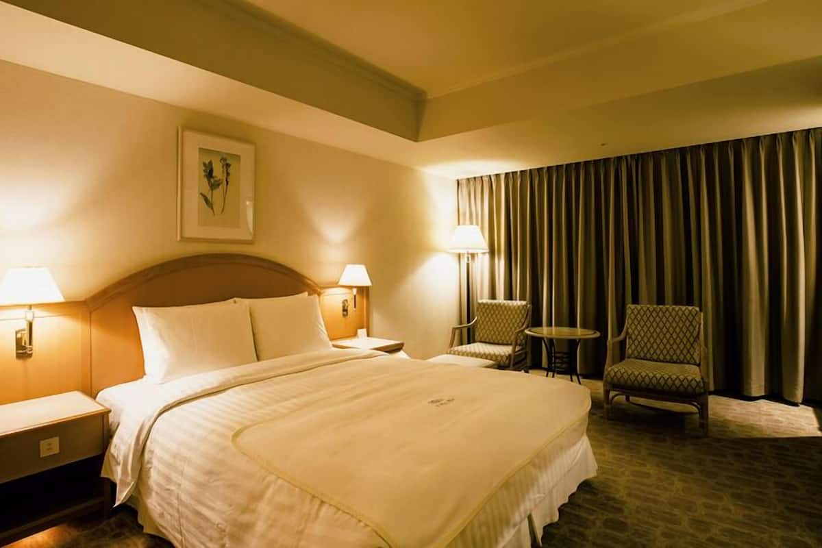Une chambre d’hôtel avec un grand lit, deux tables de chevet, une chaise, une petite table et une grande fenêtre avec des rideaux.