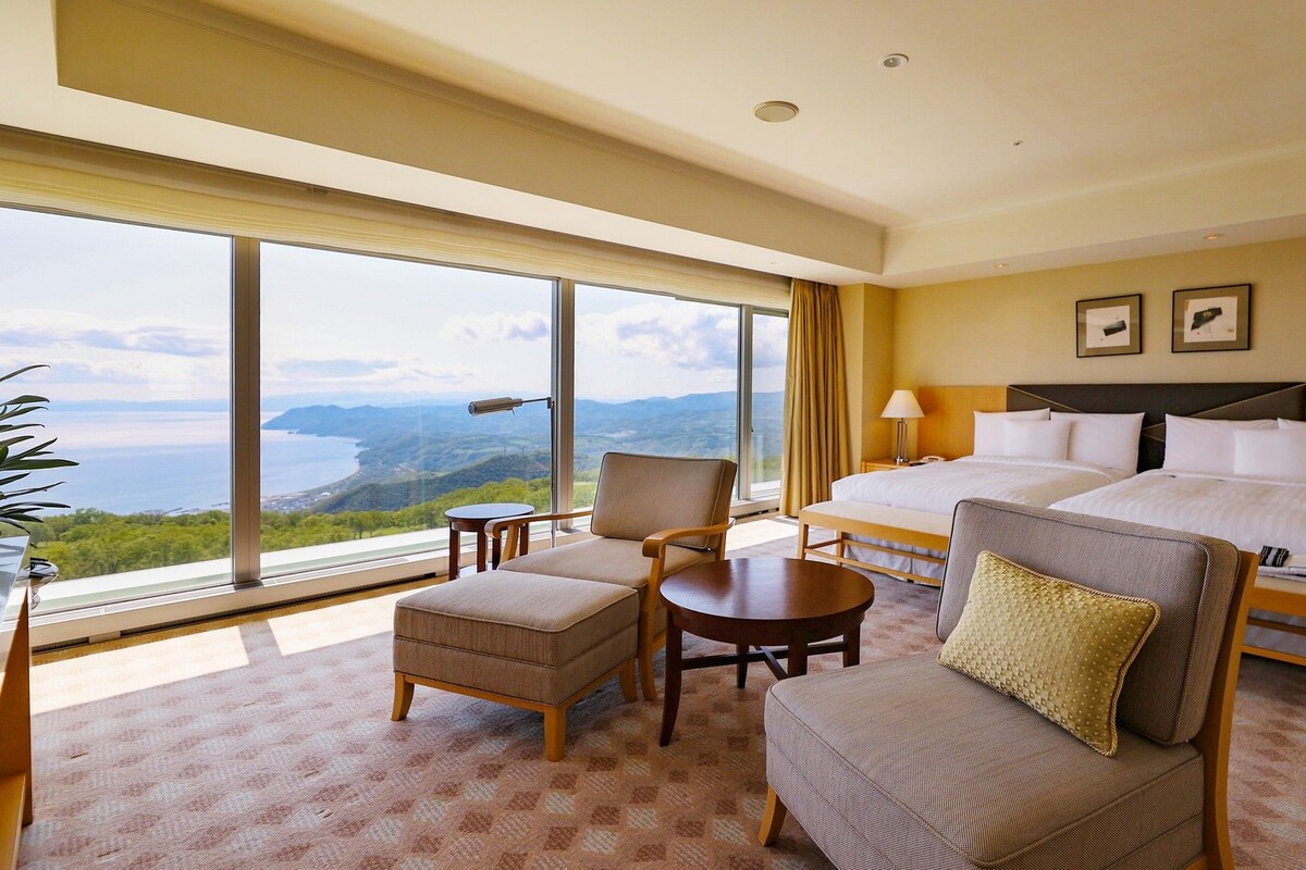 A hotel room with a large window, two beds, a chair, a small table, and a nightstand.