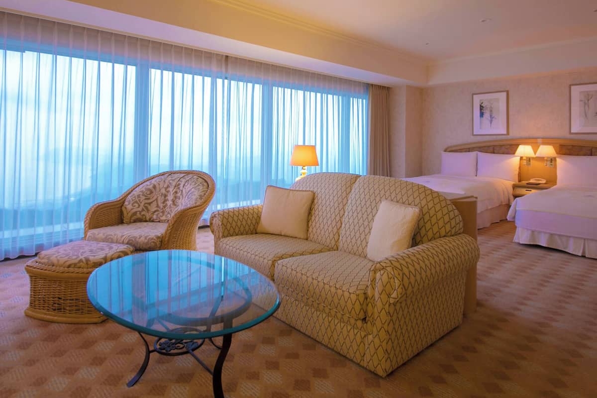 Une chambre d’hôtel avec un canapé, une table basse en verre, un lit, une table de chevet et une lampe.