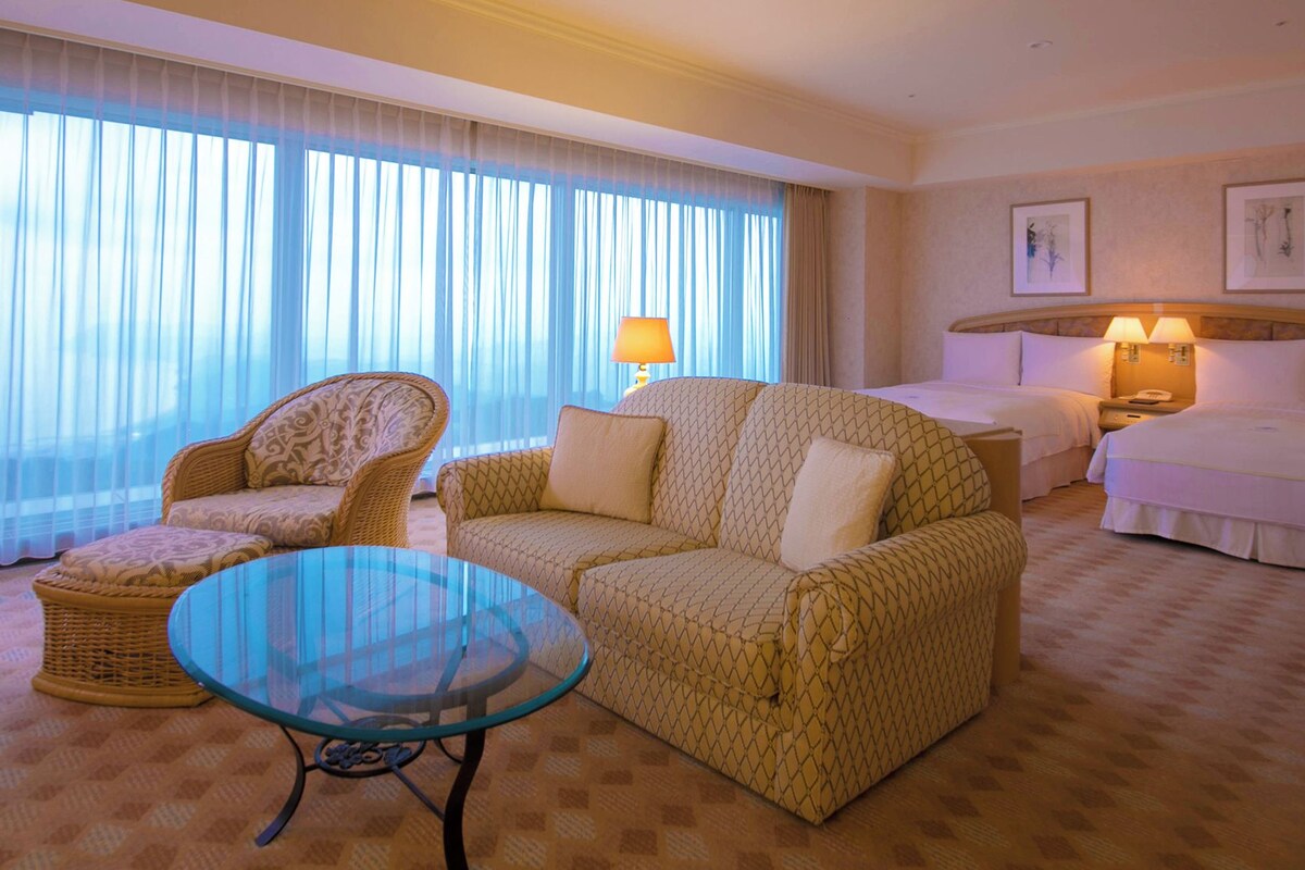 Una habitación de hotel con sofá, una mesa de centro de vidrio, cama, mesita de noche y lámpara.