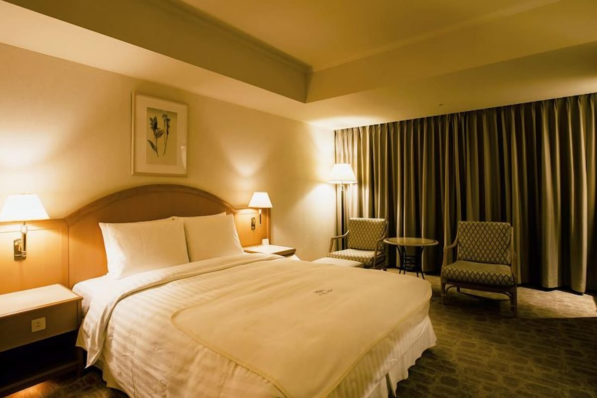 Ein Hotelzimmer mit einem großen Bett, zwei Nachttischen, einem Sessel, einem kleinen Tisch und einem großen Fenster mit Vorhängen.