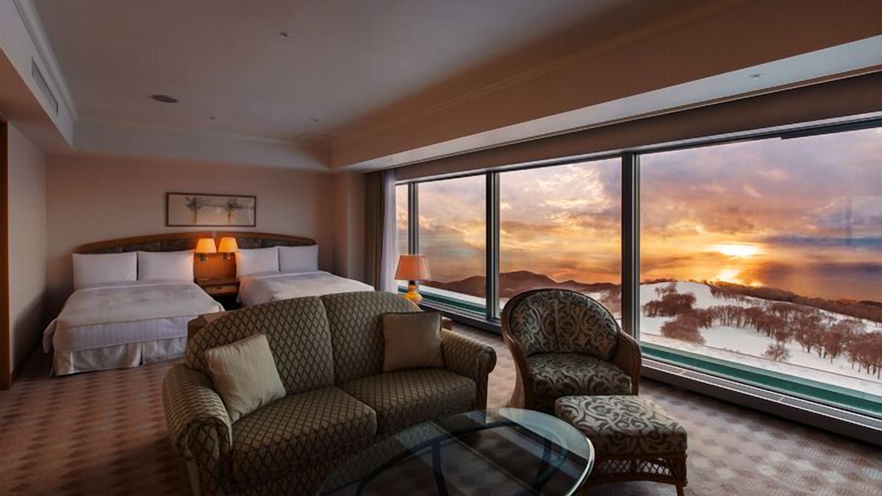 Ein Hotelzimmer mit Bett, Sofa, Sessel und einem großen Fenster mit Blick auf einen Sonnenuntergang über einer Berglandschaft.
