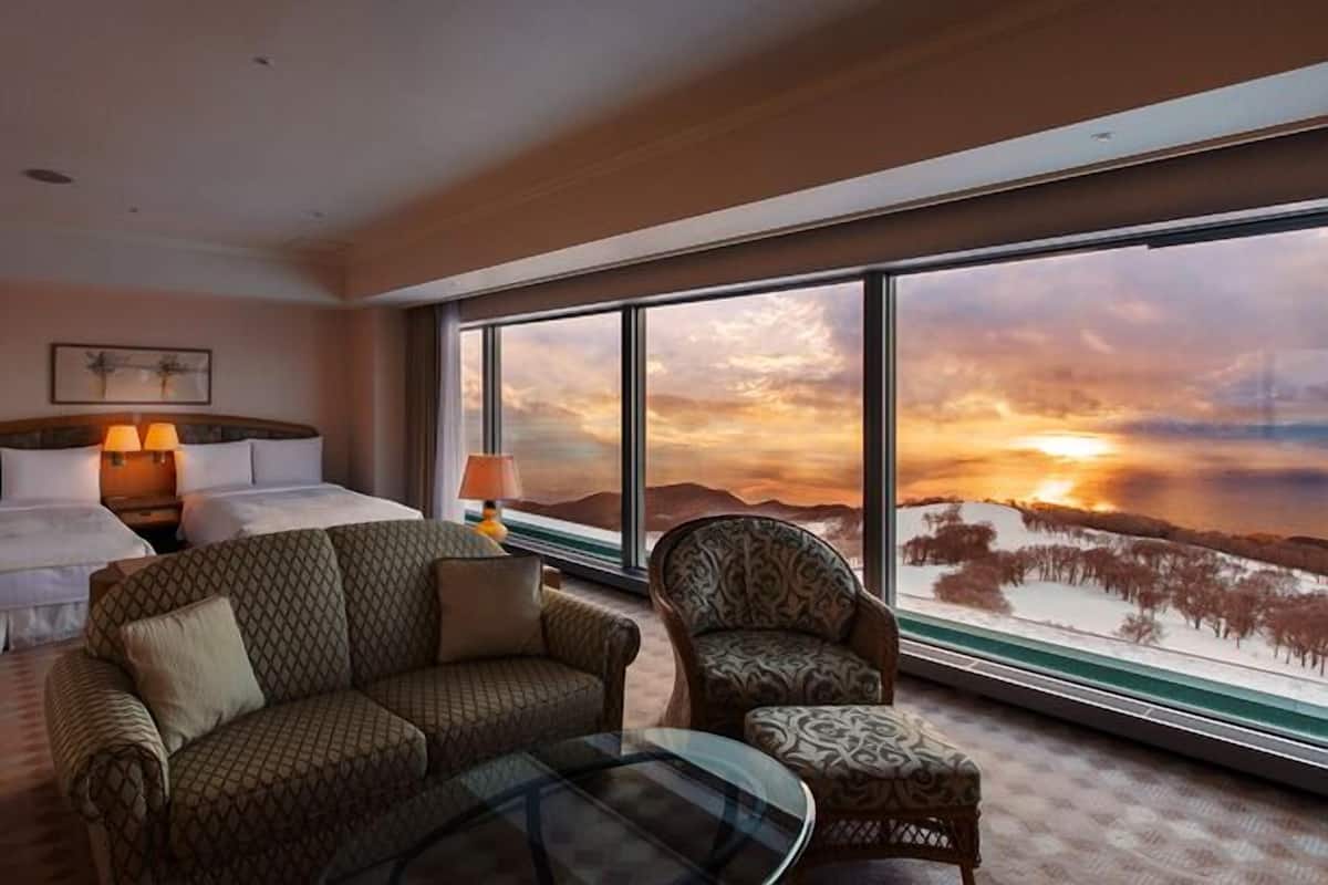 Une chambre d’hôtel avec un lit, un canapé, un fauteuil et une grande fenêtre donnant sur un paysage montagneux et offrant une vue sur le coucher du soleil.